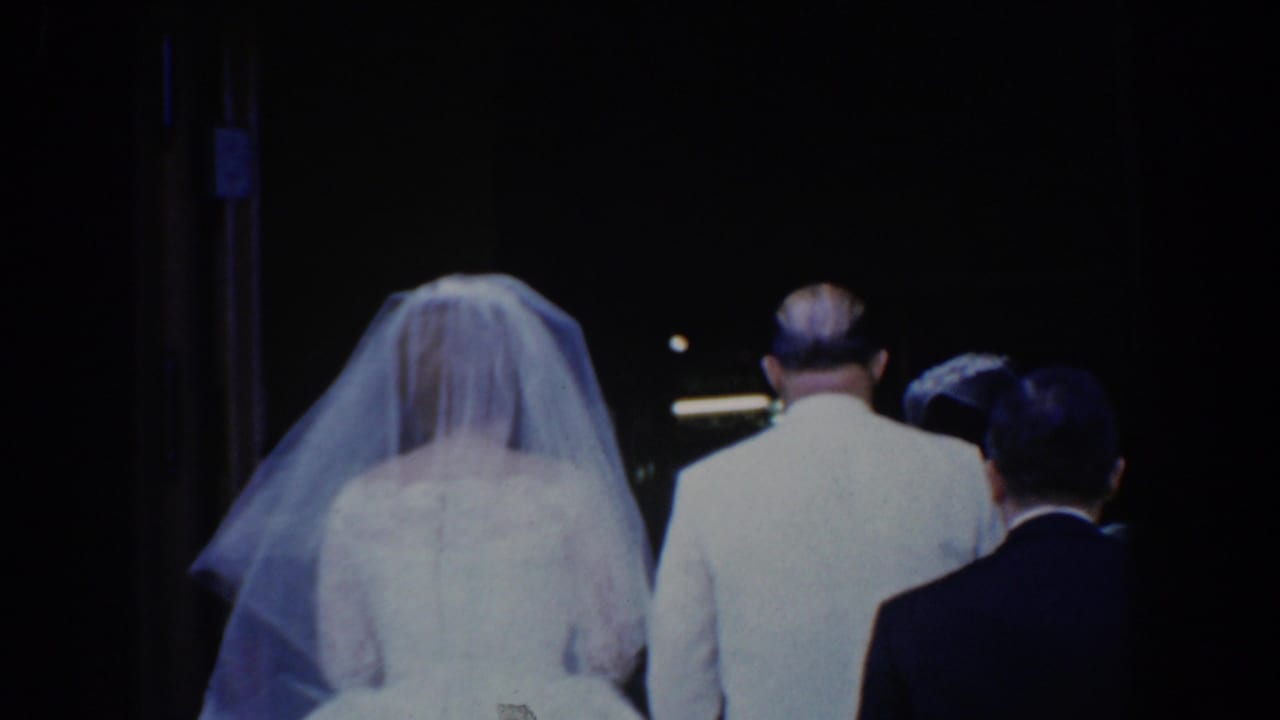 Bride in a white veil walks beside a man in a light suit with other attendants ahead.