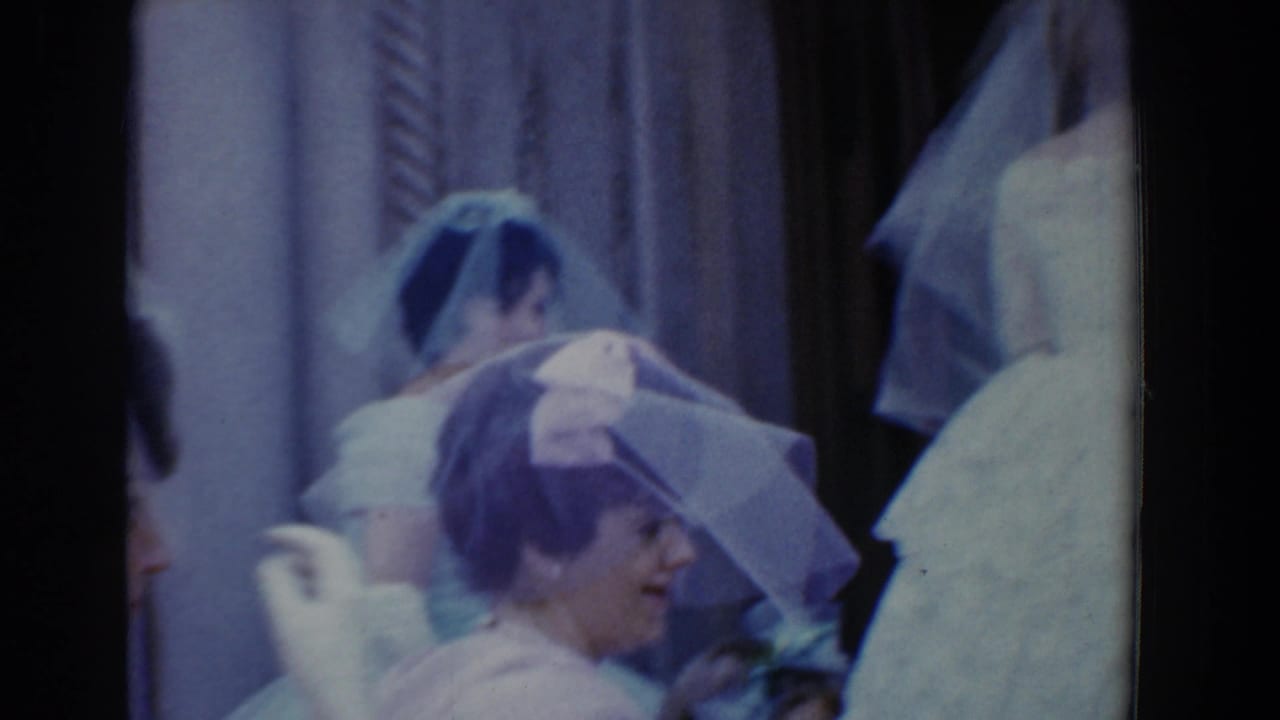 Bridesmaids or wedding attendants in white dresses and hats stand close together near curtains in the 1960 Bel Air wedding footage.