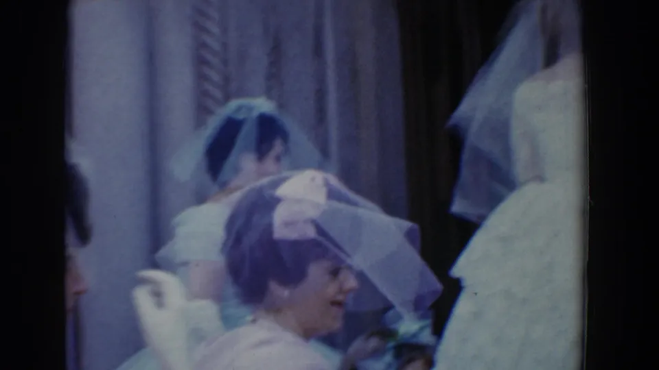 Bridesmaids or wedding attendants in white dresses and hats stand close together near curtains in the 1960 Bel Air wedding footage.