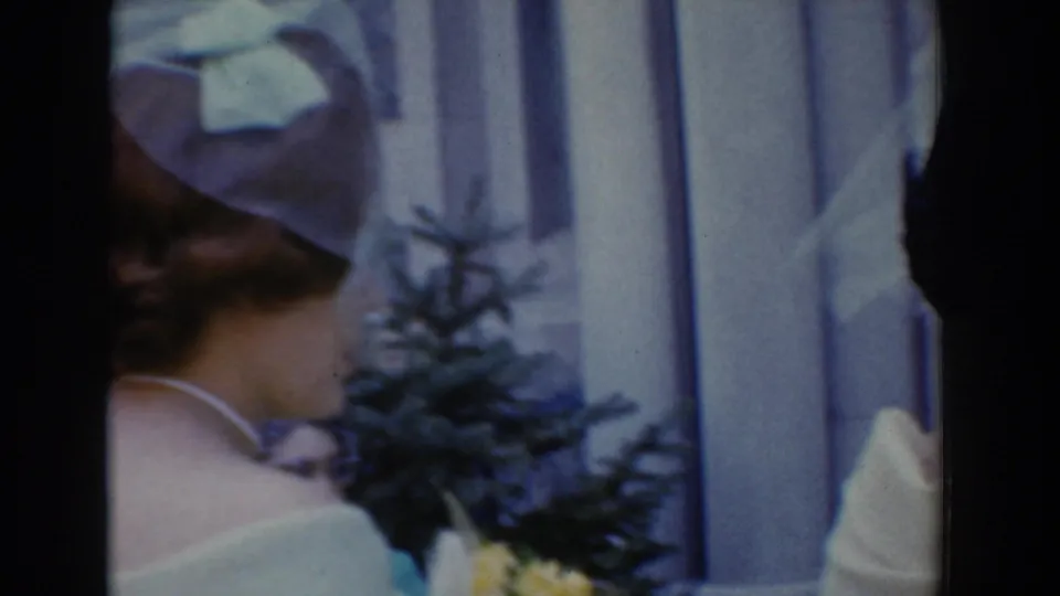 A close, soft 8mm view shows a wedding attendant in a white structured hat and dress near curtains and flowers.