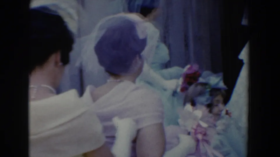 Wedding attendants in white dresses and veiled hats cluster together, one near a bouquet, in the Bel Air home-movie wedding scene.