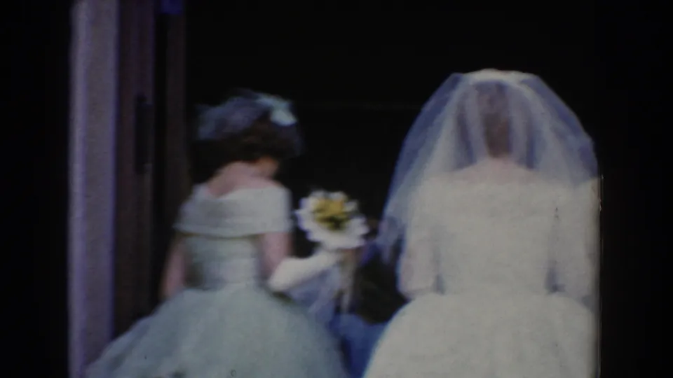 In 1960 Bel Air, Maryland, a veiled bride and bridesmaid approach a dim church doorway in 8mm home-movie footage.