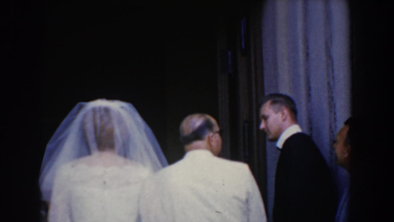 In 1960 Bel Air, Maryland home-movie footage, a veiled bride stands with her father near a dim doorway as a man in a dark suit smiles nearby.