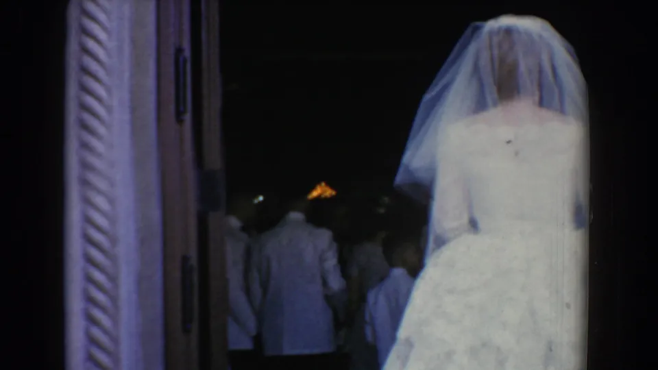 A veiled bride in a white gown stands near the open doorway as several figures move farther inside the dark room during the wedding sequence.