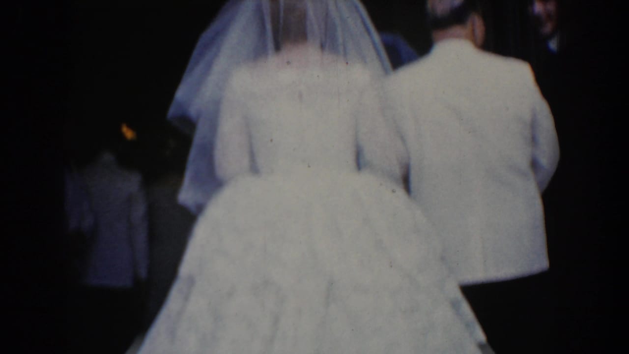 In 1960 Bel Air, Maryland footage, a bride in a white gown and veil walks from behind with a male escort through a dim venue.