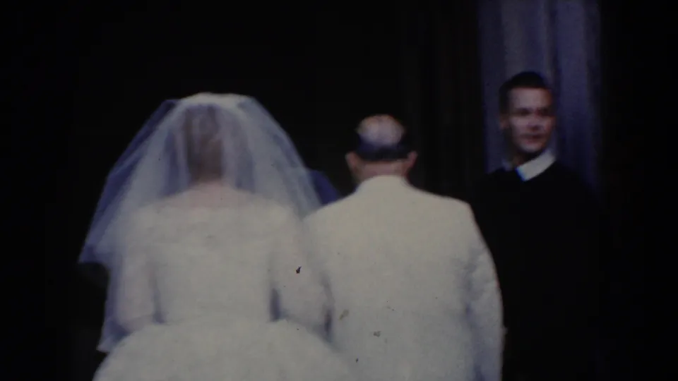 The bride and escort approach a doorway in 1960 Bel Air footage, with a suited man visible ahead in the shadows.