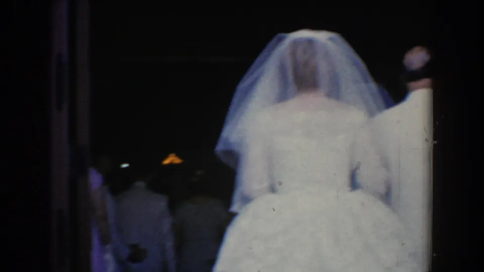 Rear view of the bride in a veil and white gown beside her escort in a dim 1960 Bel Air wedding interior.