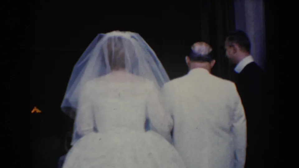 In Bel Air, Maryland, a veiled bride walks arm-in-arm with a man in a light suit as they leave a dim church doorway in 1960 home-movie footage.