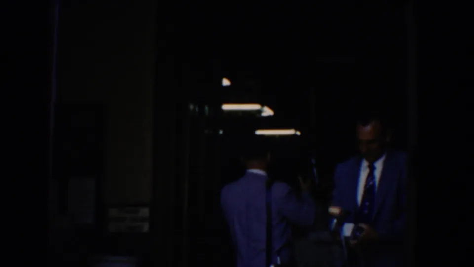 Inside a dim hallway, formally dressed men stand near overhead lights, including one man who appears to hold a camera.