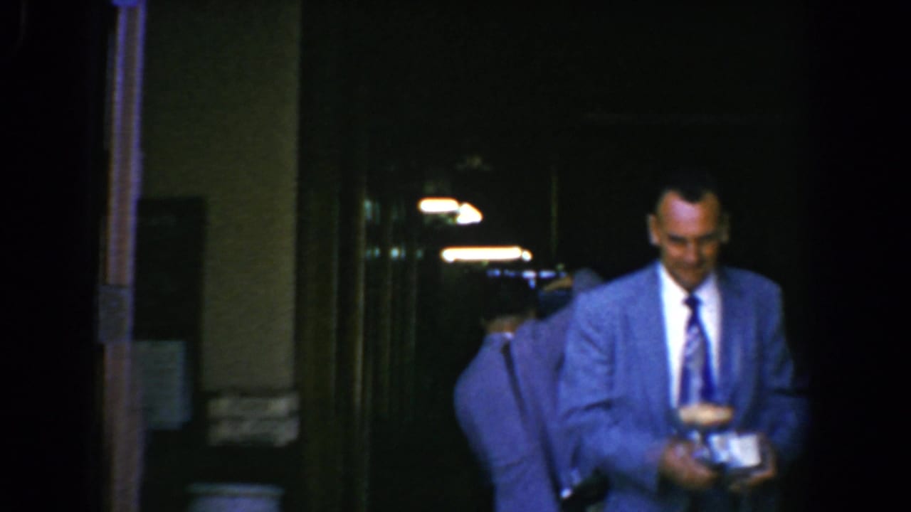 Suited man holding a camera accessory near a dark doorway at night.
