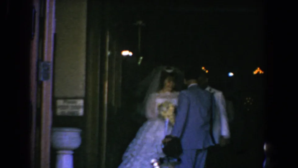A bride in a white gown and veil stands with suited men near a doorway in the 1960 Bel Air, Maryland wedding footage.