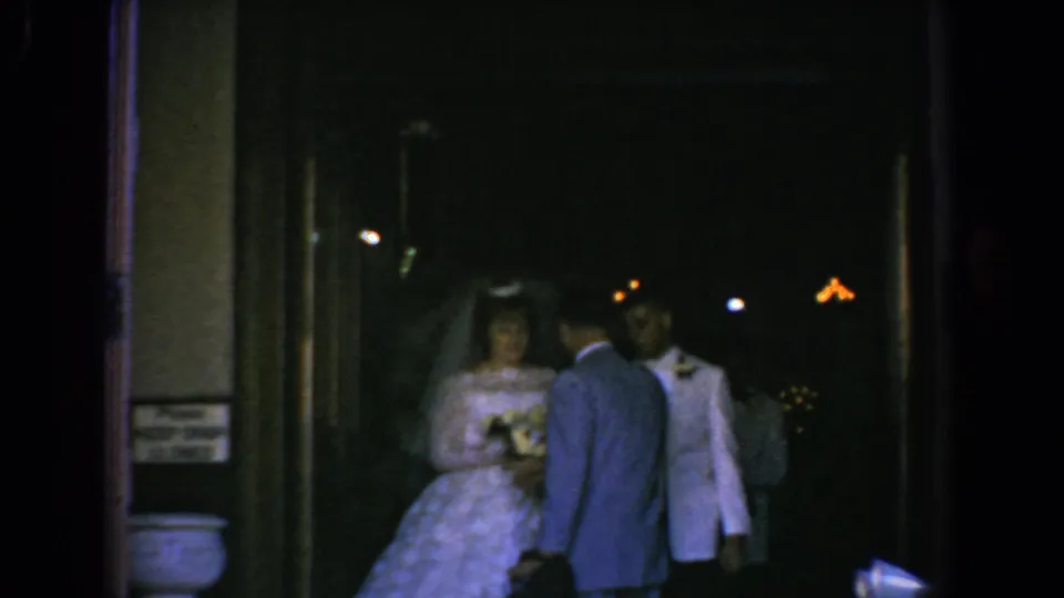 The bride holds flowers while two suited men stand close by near the dark entrance in the Bel Air wedding home movie.