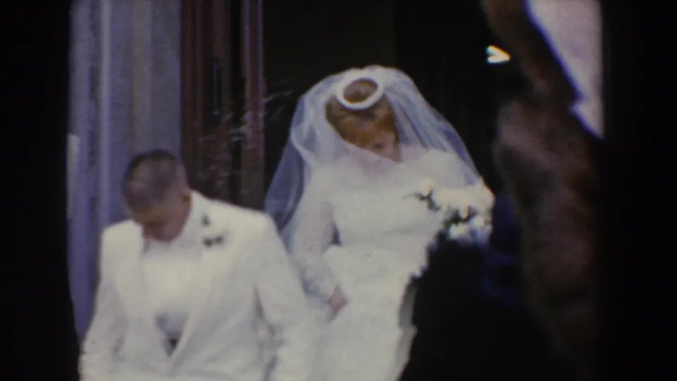 1960 Bel Air, Maryland wedding exit with bride carrying a bouquet beside the groom as the handheld camera is partly blocked by guests.