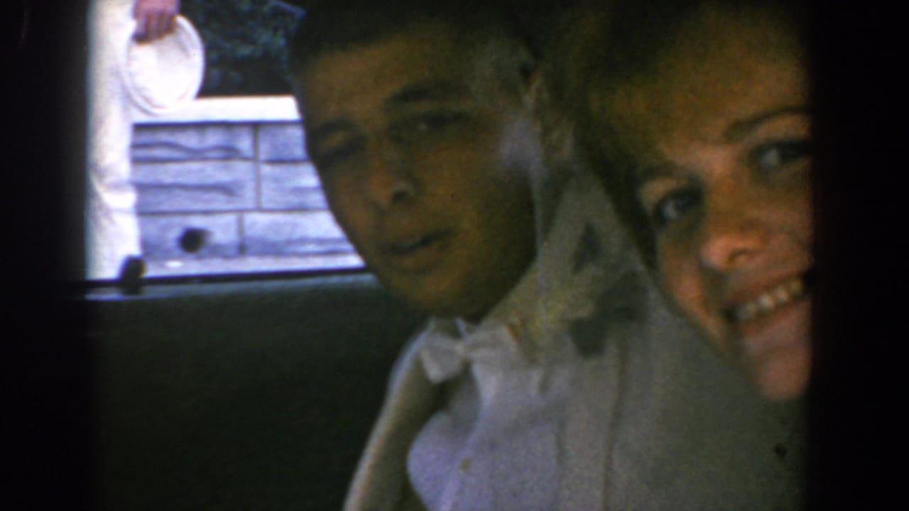 The couple remains close to the open car window, with the woman in white and the man in a light suit facing the camera.