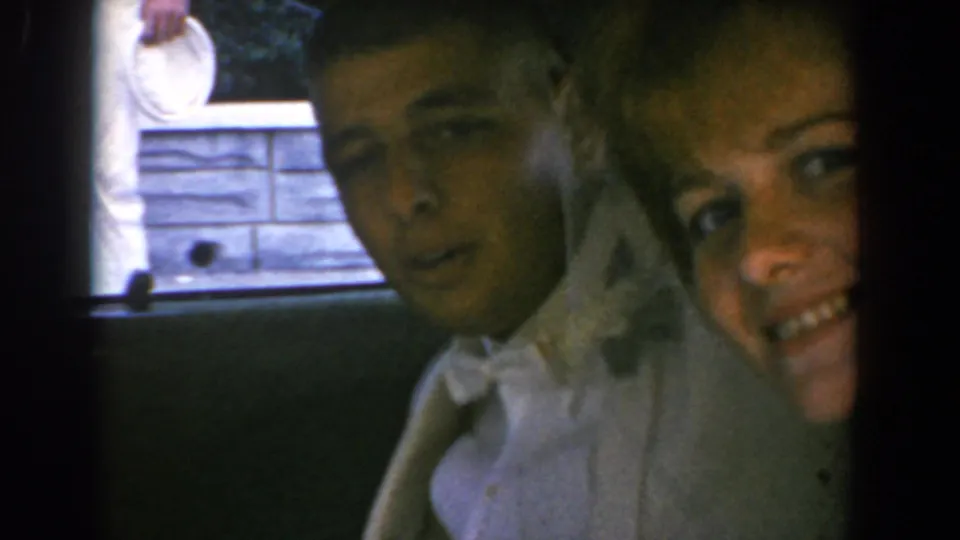 The couple remains close to the open car window, with the woman in white and the man in a light suit facing the camera.