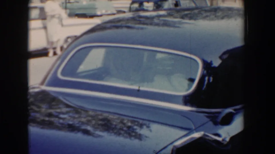 A close 8mm view of the black car's rear window and curved bodywork, with parked vehicles and bright daylight behind it.