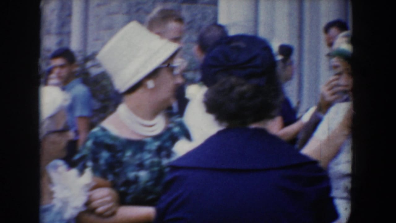Formally dressed adults gather outside a stone building, including a woman in a light hat and guests in dark clothing.