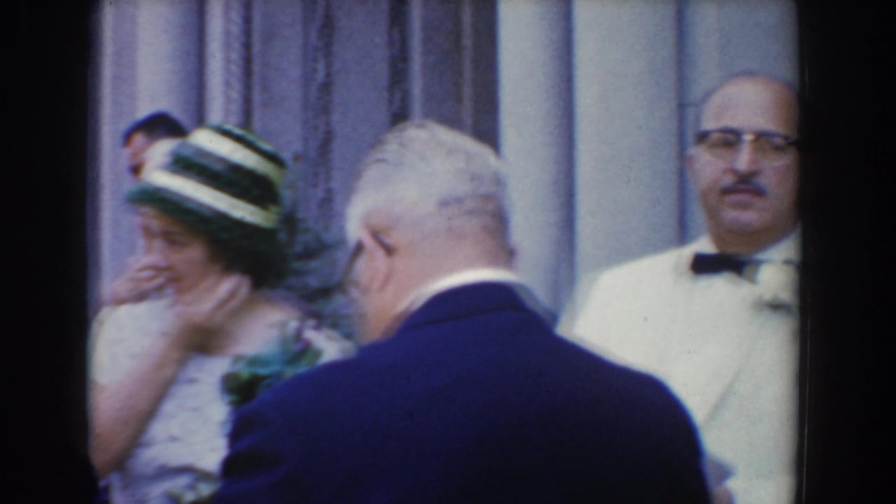 A woman in a striped hat stands near an older man in a dark suit and a man in a white jacket with bow tie.