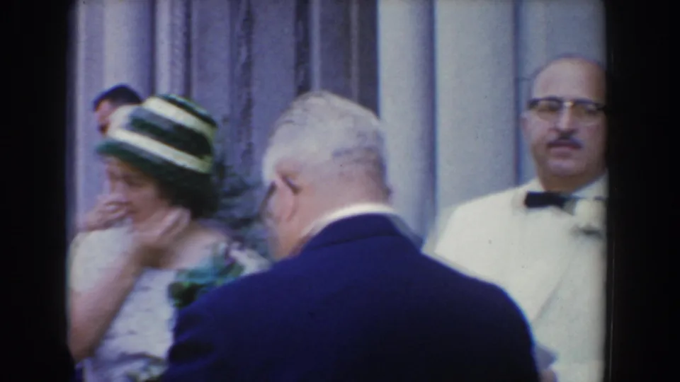 A woman in a striped hat and corsage stands among formally dressed adults near columns in Bel Air, Maryland, 1960.