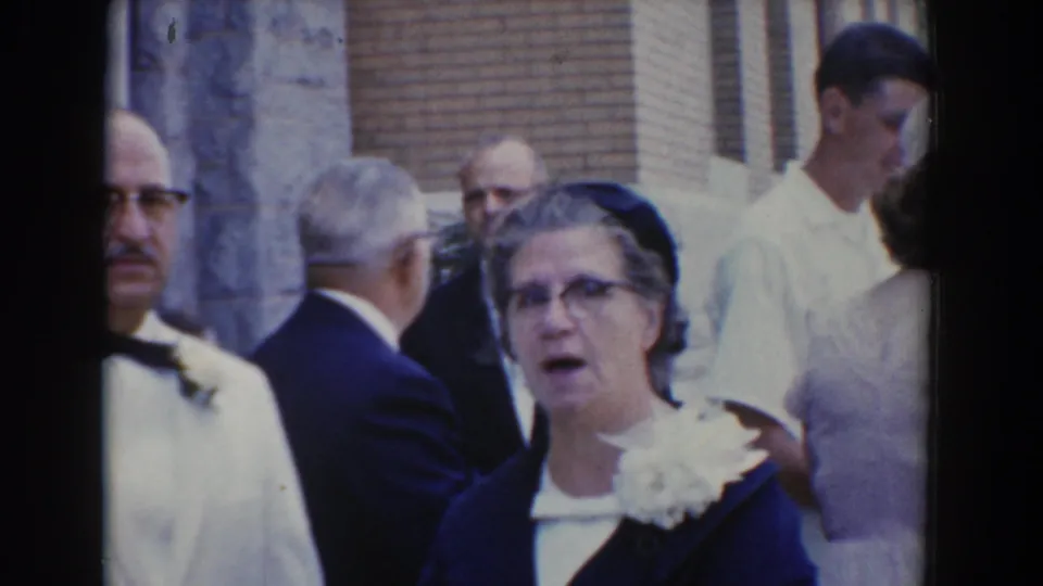 Adults in formal dress stand near a brick and stone exterior in 1960 Bel Air, with faces and corsages visible in the crowd.