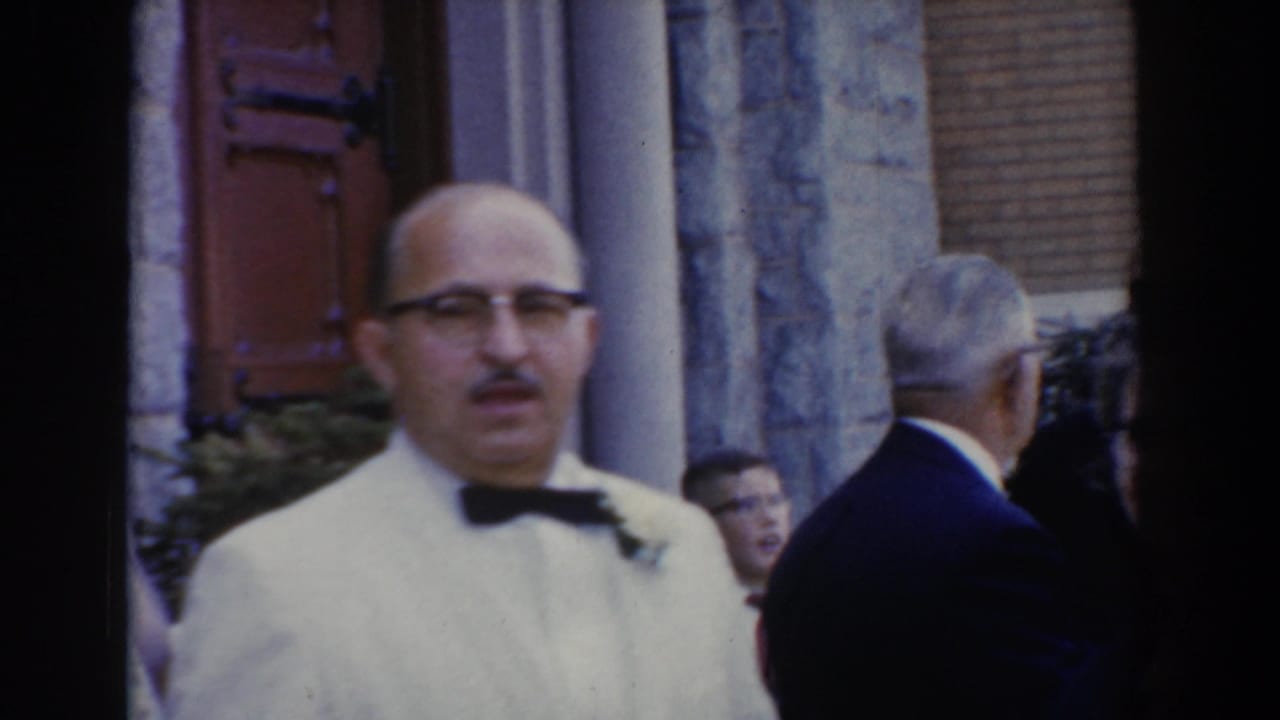 In 1960 Bel Air, Maryland, a man in a white tuxedo and glasses faces the camera outside a stone building during a wedding gathering.