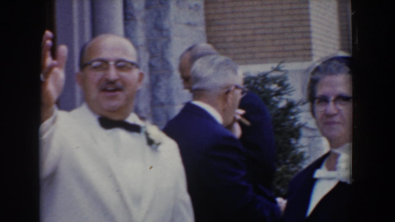 Man in a white tuxedo waves while wedding guests stand nearby.