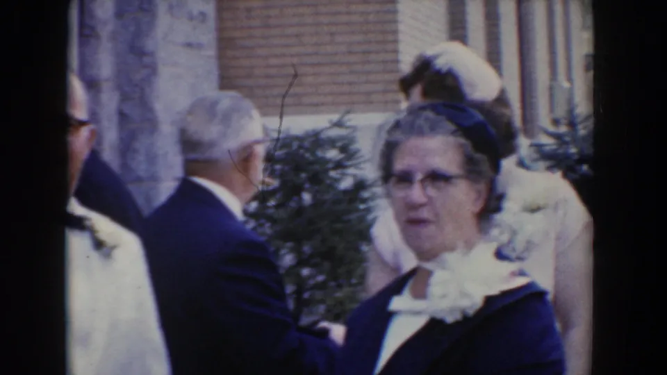 A woman in a dark outfit and hat looks toward the camera as wedding guests move outside the stone building in Bel Air, Maryland.