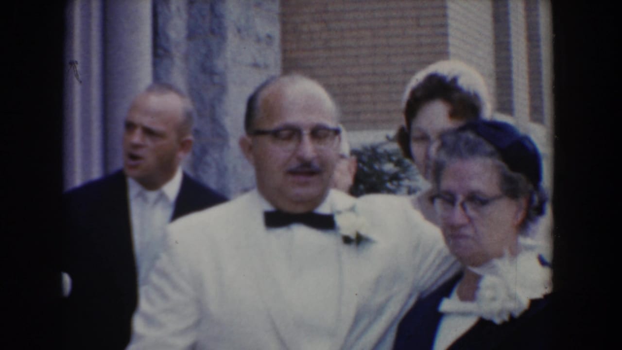 Older man in a white jacket stands with an older woman at a formal gathering.