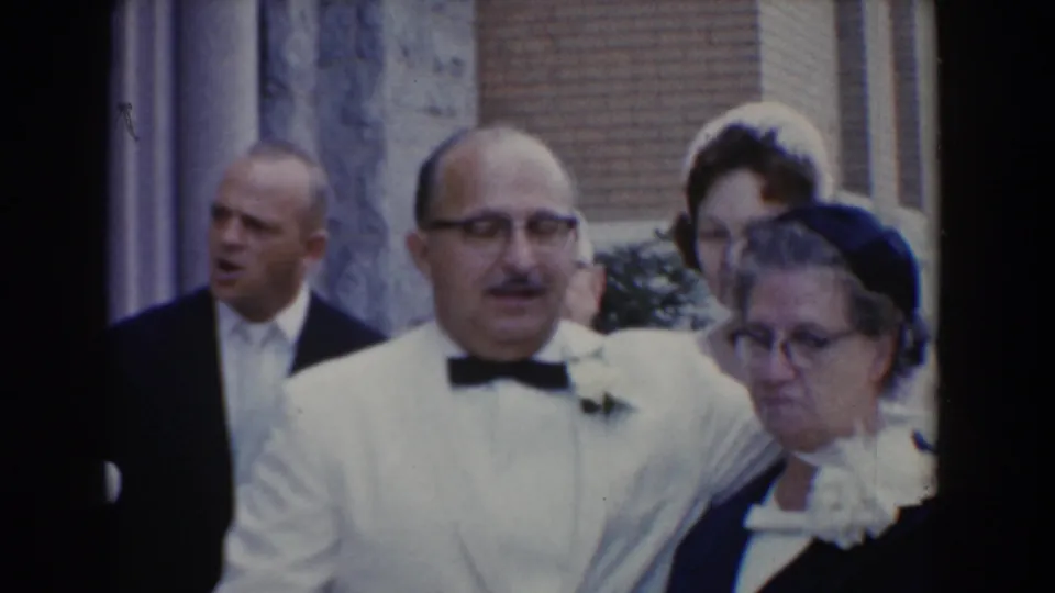 1960 Bel Air, Maryland frame showing an older man in a white dinner jacket beside an older woman in a dark dress during a formal outdoor event.