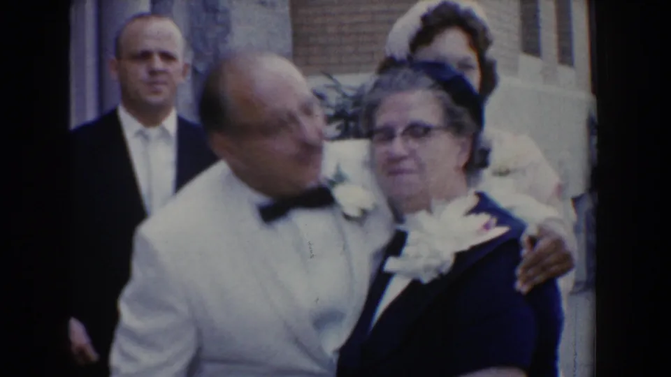 1960 Bel Air, Maryland frame showing an older man in a white dinner jacket leaning toward an older woman in a dark dress as guests stand nearby.