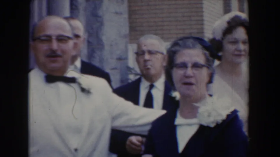 1960 Bel Air, Maryland frame showing an older couple at a formal outdoor gathering, with the man in a white jacket and the woman in a dark dress.