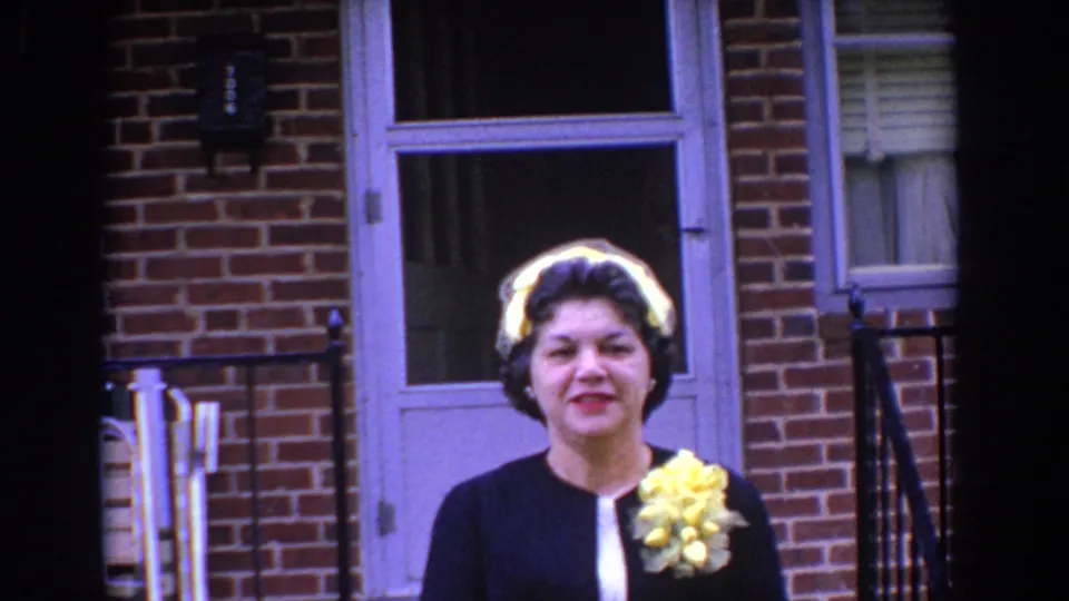 A close view shows the woman facing the camera outside the Bel Air brick home, with a yellow corsage, white gloves, and formal black suit.