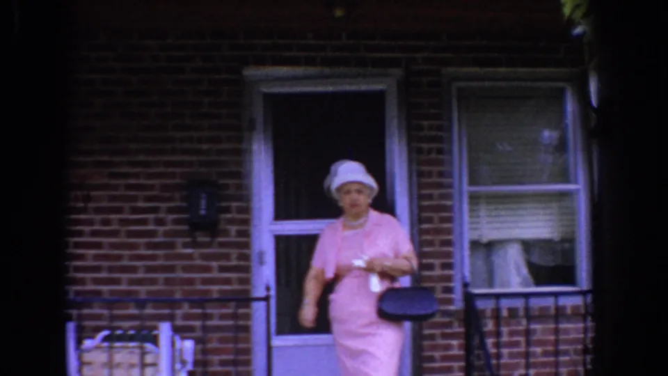 A second woman in a pink dress and light hat appears at the doorway of the Bel Air residence, holding a dark handbag.