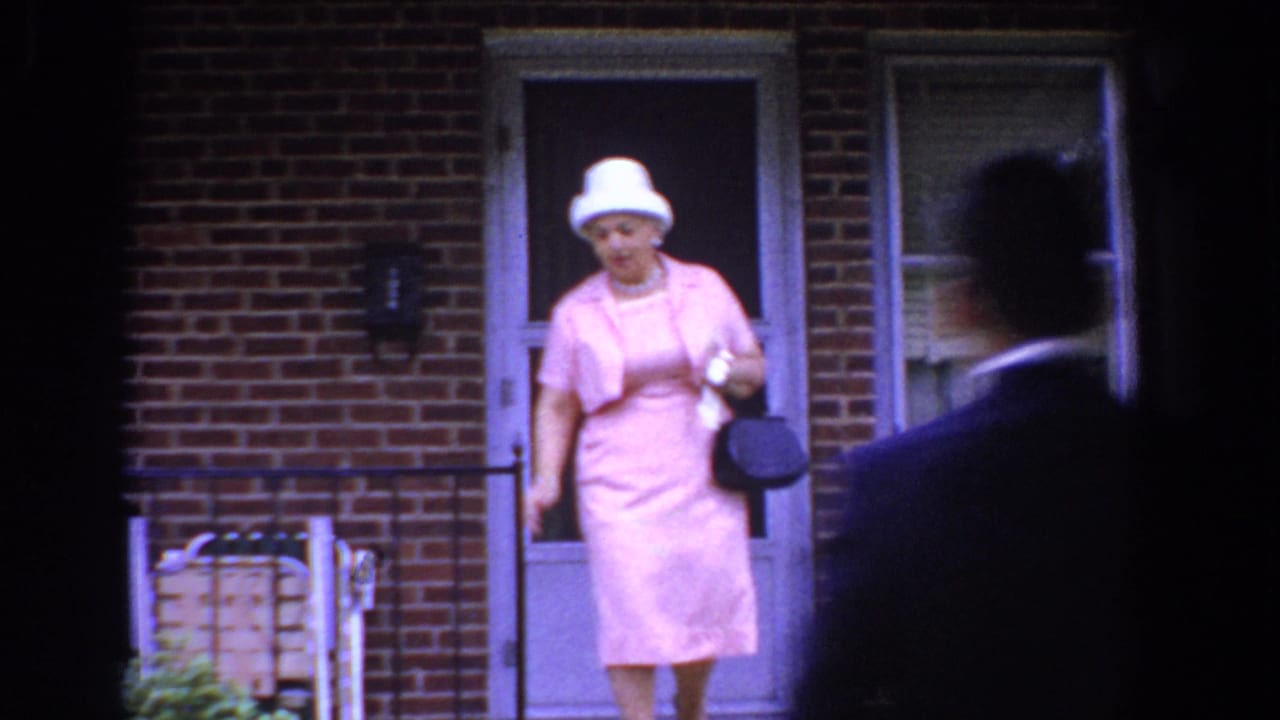 Woman in a pink dress and white hat steps from a brick building doorway as a suited man passes in front.