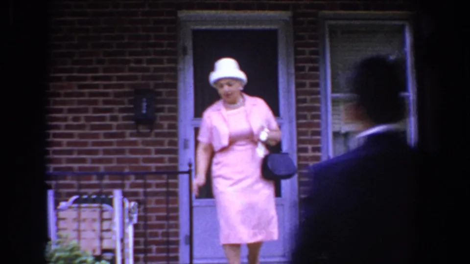 In Bel Air, Maryland, 1960, a woman in a pink dress and white hat exits a brick residence while a suited man crosses the foreground.