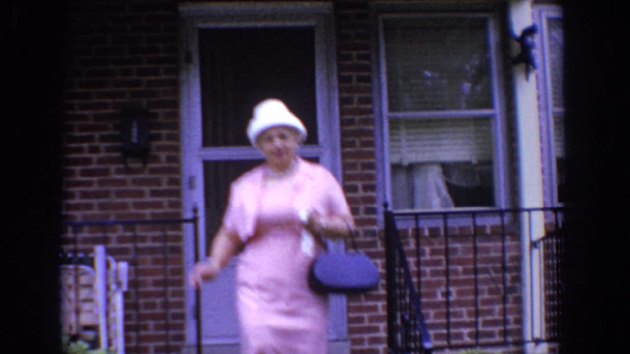 Woman in pink walks along a brick building entrance carrying a dark handbag and small light item.