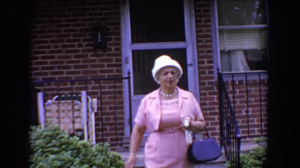 In the 1960 Bel Air home-movie footage, the woman approaches the camera, showing her white hat, necklace, handbag, and pink dress.