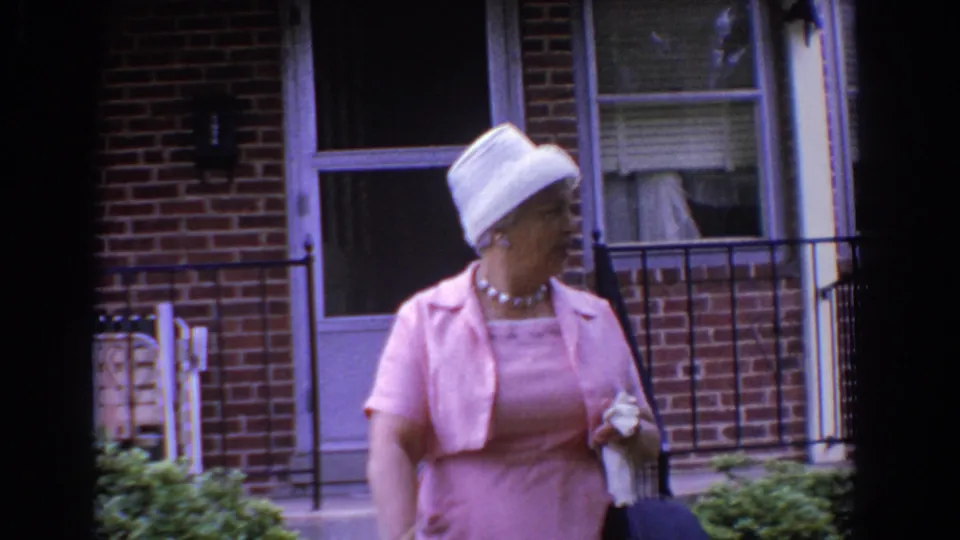 The woman turns her head near the brick residence entrance, holding her handbag and light item as the 8mm frame softens with motion.