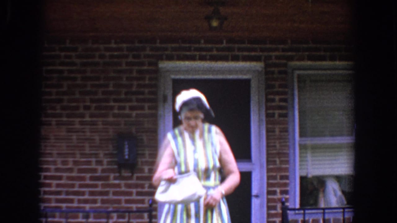 In Bel Air, Maryland, around 1960, a woman in a striped dress and white hat stands by a brick home doorway holding a handbag.