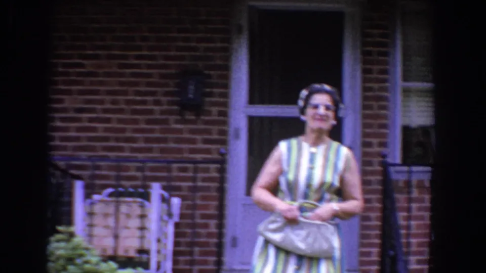 A woman in 1960 Bel Air, Maryland, smiles near porch steps while holding a light handbag in front of a brick residential exterior.