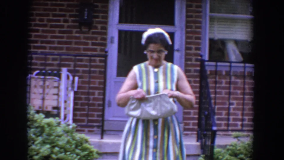 The woman looks down and adjusts her light handbag beside the porch steps of a brick home in Bel Air, Maryland, around 1960.