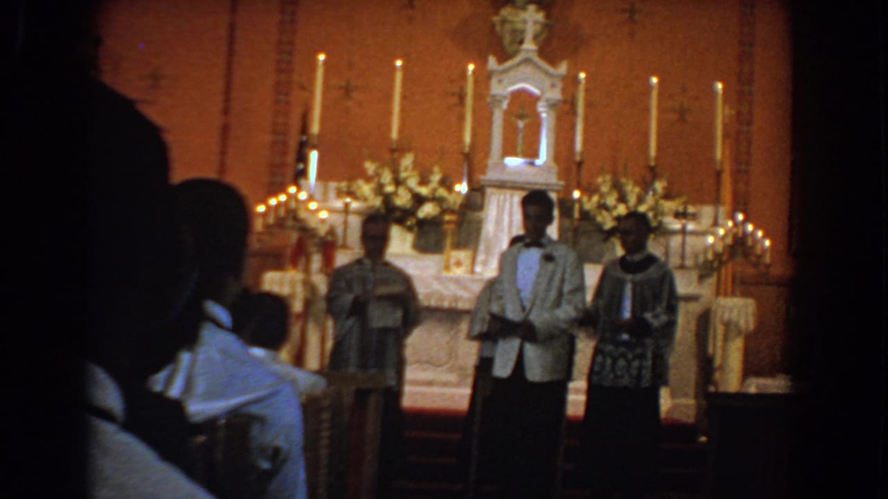 1960 Bel Air, Maryland home-movie frame showing a formally dressed man before a candlelit church altar with clergy and seated attendees.