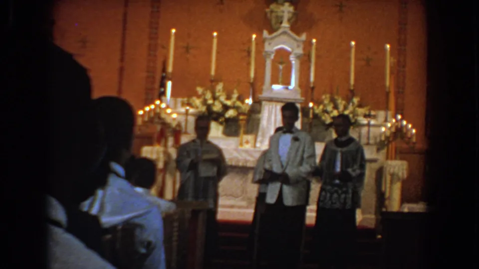 1960 Bel Air, Maryland home-movie frame showing a formally dressed man before a candlelit church altar with clergy and seated attendees.