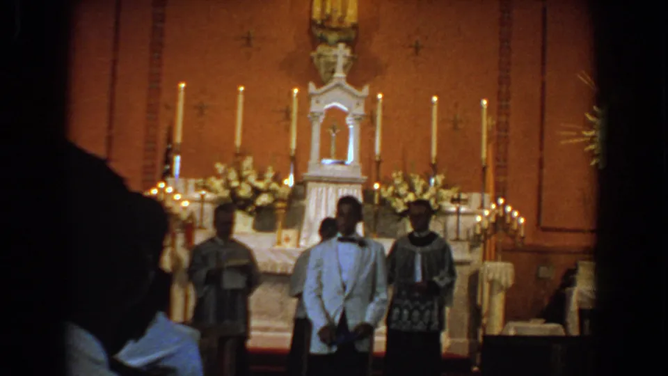 A formally dressed man stands before clergy and a decorated altar in this 1960 Bel Air, Maryland church ceremony frame.