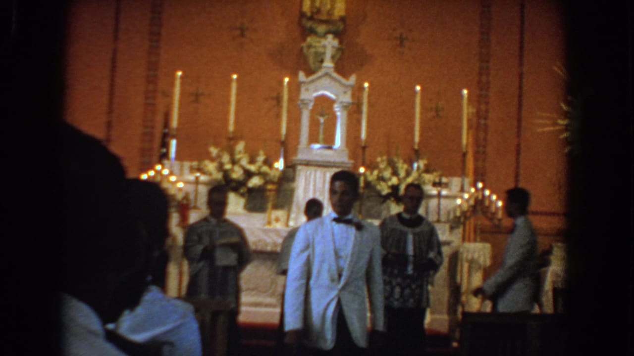 Young man in a white tuxedo jacket stands near clergy at a candlelit church altar.