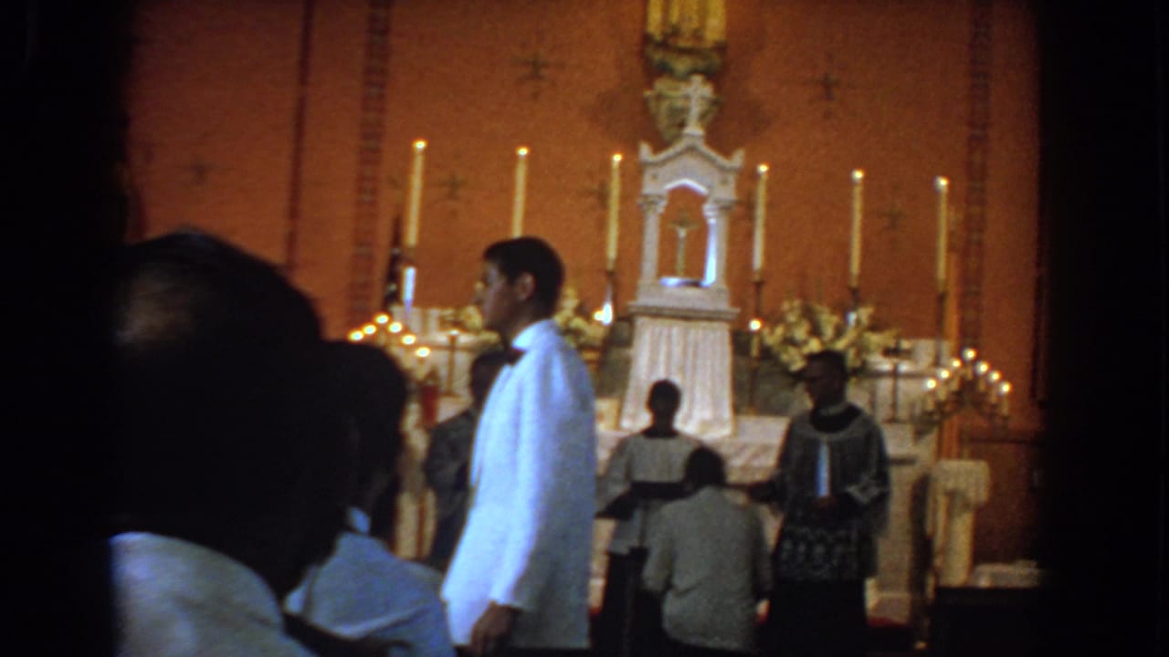 Young man in white formal jacket walks across the front of a candlelit church altar.
