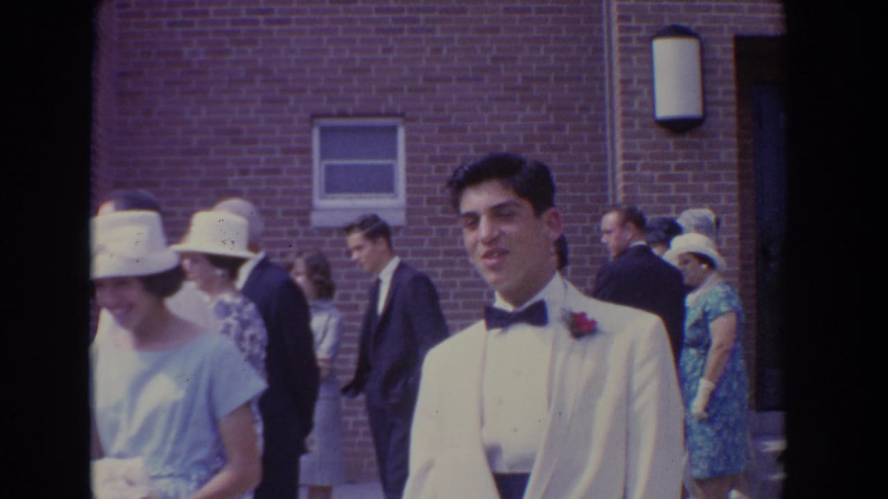 Groom in a white tuxedo jacket near women in hats outside a brick church.