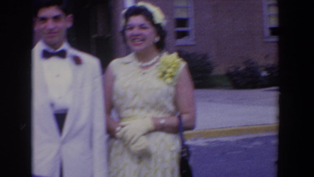 Man in a white dinner jacket and woman in a pale dress stand near a brick building.