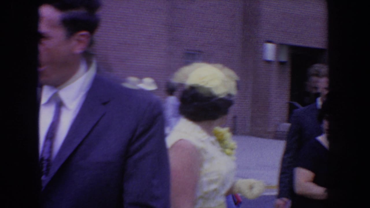 Man in a dark suit stands beside a woman in a pale dress and hat near a crowd.
