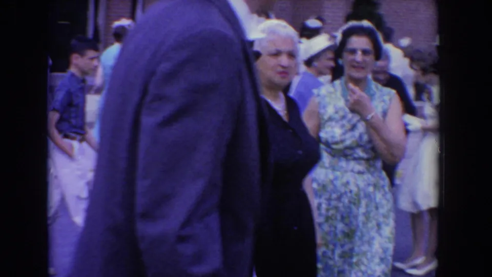 Adults in mid-century formal clothing gather outdoors in Bel Air, Maryland, with a suited man crossing the foreground.